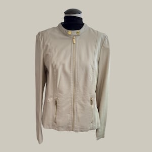 May include: A tan faux leather jacket with a stand-up collar and gold-colored zipper and hardware. The jacket has two zippered pockets and is displayed on a mannequin with a black turtleneck.