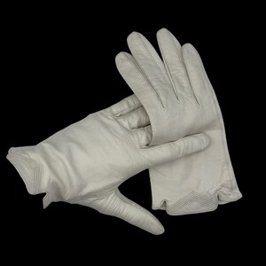 May include: A pair of off-white leather gloves. The gloves are shown against a black background. The gloves have a classic design with a decorative stitched cuff.