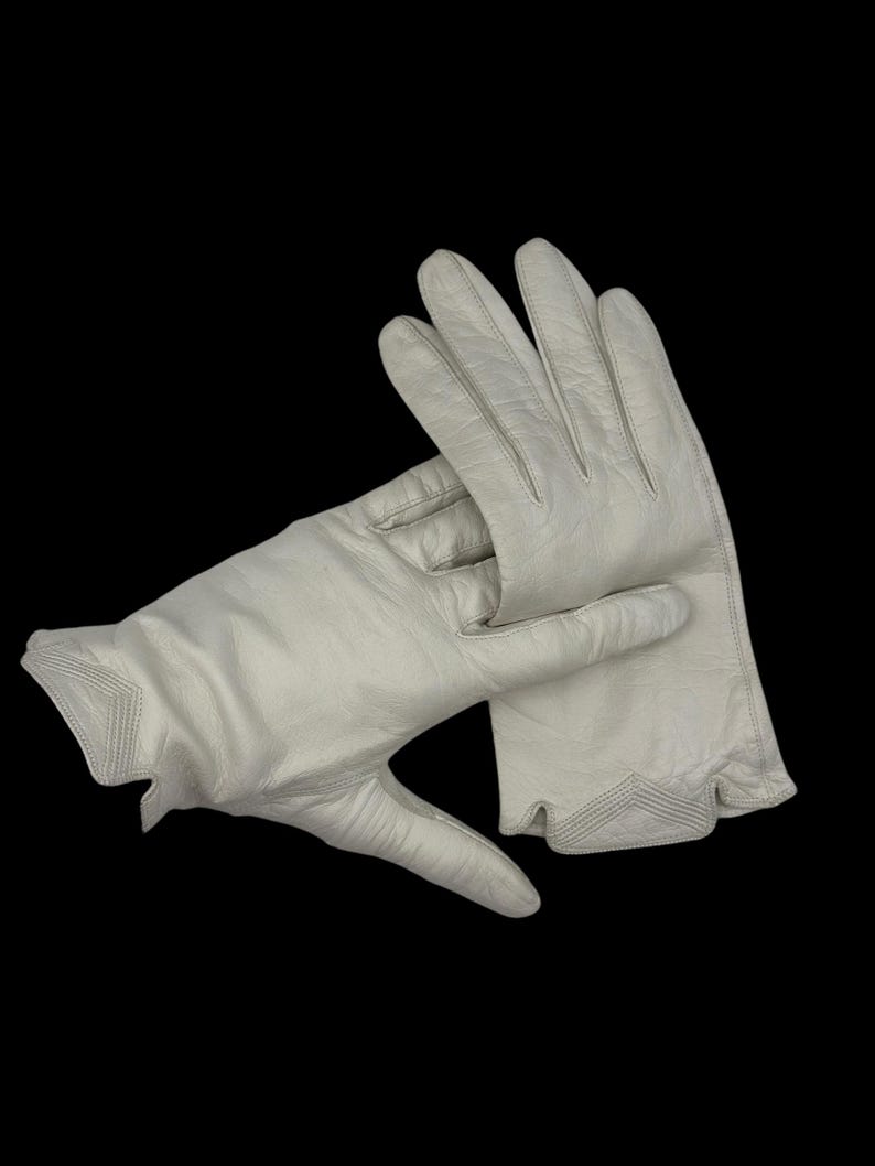 May include: A pair of off-white leather gloves. The gloves are shown against a black background. The gloves have a classic design with a decorative stitched cuff.