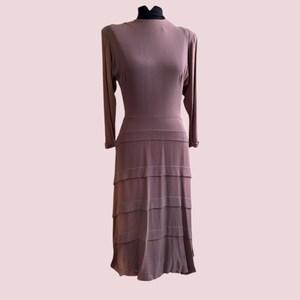 Puede incluir: Vestido color malva con mangas largas y falda escalonada. El vestido tiene un cuello redondo y se exhibe en un maniquí. El vestido parece estar hecho de una tela suave y fluida, y es de estilo vintage.