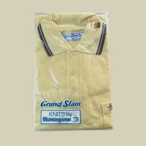 May include: A new, yellow Grand Slam polo shirt, still in its original packaging. The shirt has a classic collar with brown stripes and a small penguin logo. The packaging includes the brand name "Grand Slam" and "KNITS by Munsingwear".