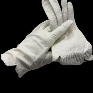 May include: A pair of white leather gloves with a textured surface. The gloves have a scalloped edge and decorative stitching on the back of the hand. The gloves are shown against a black background.