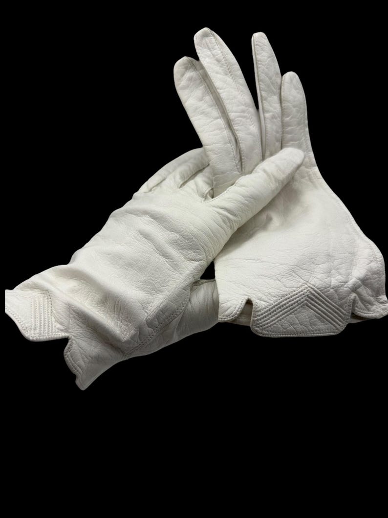May include: A pair of white leather gloves with a textured surface. The gloves have a scalloped edge and decorative stitching on the back of the hand. The gloves are shown against a black background.