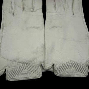 May include: A pair of off-white leather gloves. The gloves have a textured surface and feature decorative stitching at the wrist. The gloves are designed with individual finger slots and a curved edge at the wrist.