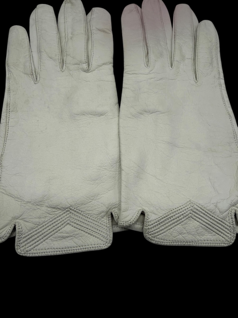 May include: A pair of off-white leather gloves. The gloves have a textured surface and feature decorative stitching at the wrist. The gloves are designed with individual finger slots and a curved edge at the wrist.