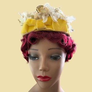 May include: A decorative hat featuring a bright yellow velvet band wrapped around the base. The top of the hat is adorned with a cluster of white fabric flowers and a single gold-toned leaf accent. The hat sits atop a mannequin head with red hair.