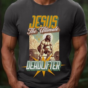 Op de afbeelding: Een donkergrijs T-shirt met een afbeelding van Jezus die een halterstang optilt. De afbeelding is in retro-stijl met gele, oranje en bruine kleuren. De tekst "Jezus De Ultieme Deadlifter" staat op het T-shirt gedrukt.