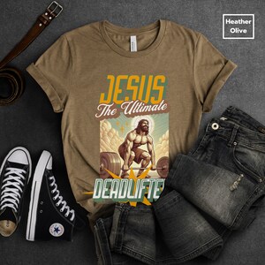 Op de afbeelding: Een bruin t-shirt met de tekst "JESUS The Ultimate Deadlifter" in vintage stijl. De afbeelding toont een cartoon van Jezus die een halterstang optilt.
