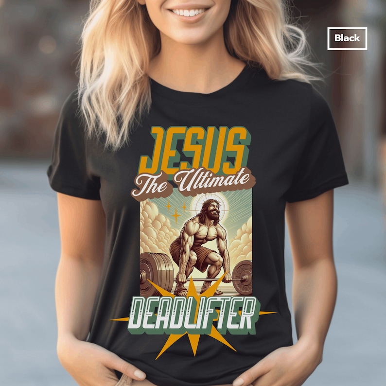 Op de afbeelding: Zwart T-shirt met een afbeelding van Jezus die gewichten heft. De tekst "Jesus The Ultimate Deadlifter" staat op het shirt gedrukt.