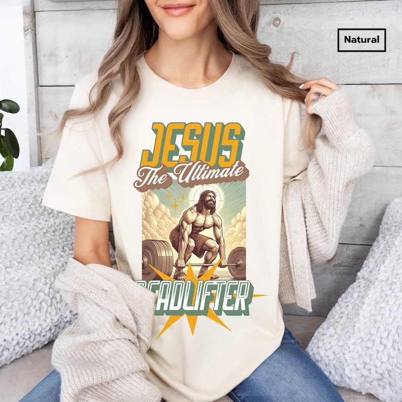 Op de afbeelding: Een beige t-shirt met een afbeelding van Jezus die gewichten heft. De afbeelding bevat de tekst "Jesus The Ultimate Deadlifter" in een retro-stijl lettertype.