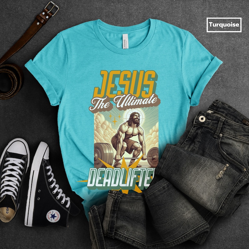 Op de afbeelding: Een turquoise T-shirt met een afbeelding van Jezus die een halterstang optilt met de tekst "Jesus The Ultimate Deadlifter".