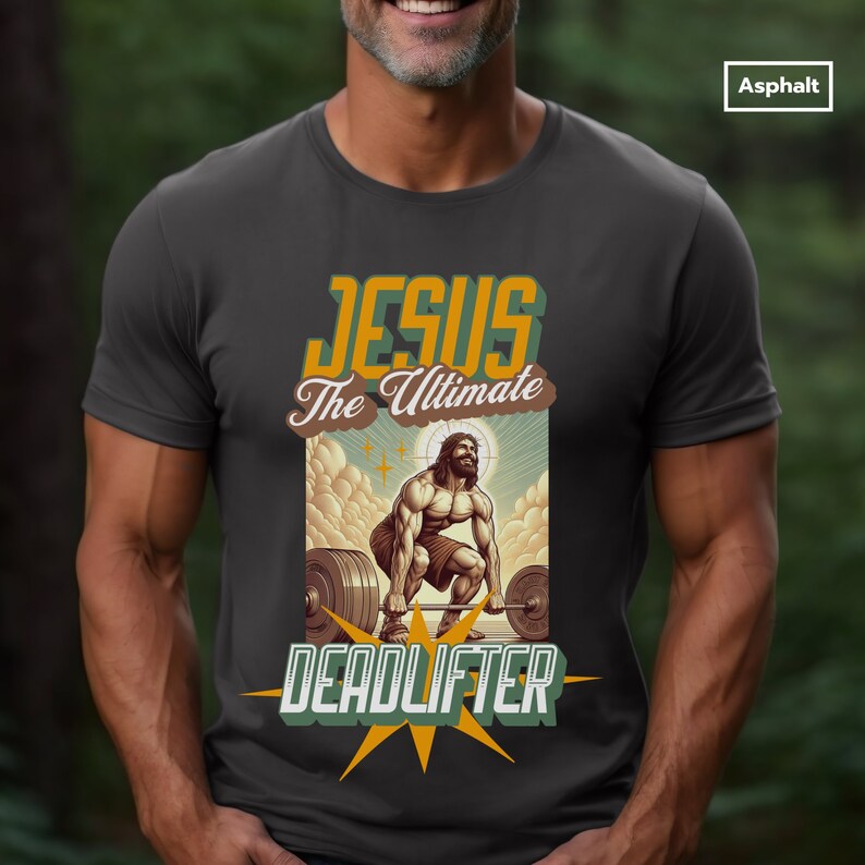 Op de afbeelding: Een donkergrijs T-shirt met een afbeelding van Jezus die een halterstang optilt. De afbeelding is in retro-stijl met gele, oranje en bruine kleuren. De tekst "Jezus De Ultieme Deadlifter" staat op het T-shirt gedrukt.