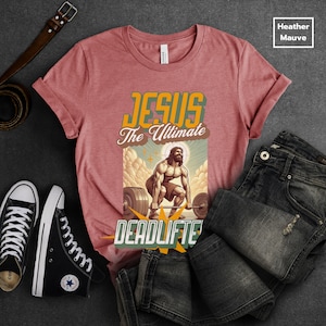 Op de afbeelding: Een heather mauve t-shirt met een afbeelding van Jezus die gewichten heft. De afbeelding zegt "Jesus The Ultimate Deadlifter".