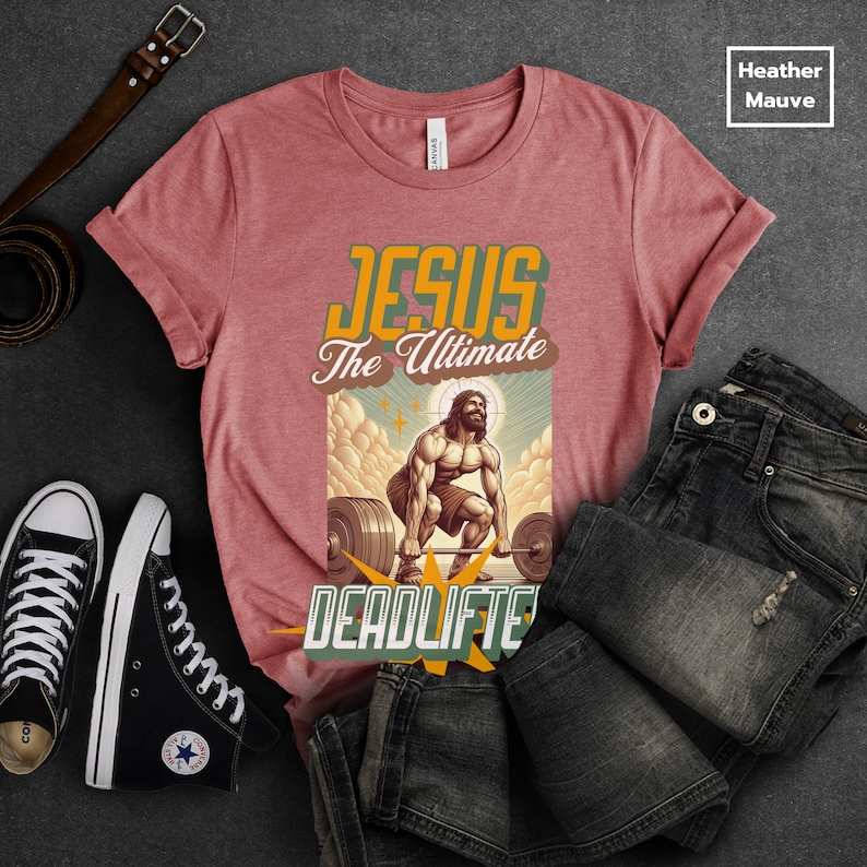 Op de afbeelding: Een heather mauve t-shirt met een afbeelding van Jezus die gewichten heft. De afbeelding zegt "Jesus The Ultimate Deadlifter".