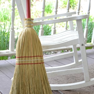 May include: A straw broom with a red handle leaning against a white rocking chair on a wooden porch.