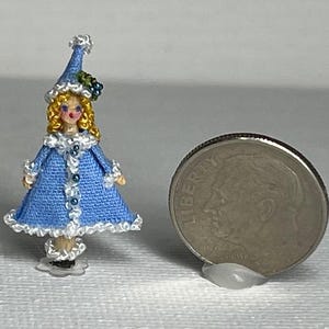 May include: A miniature doll with a blue dress and hat, accented with white trim and bead details. The doll has blonde curly hair and is next to a dime coin for size comparison. The doll is approximately 1.5 inches tall.