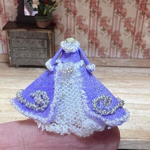 May include: Miniature lavender dress with white lace and silver bead accents. The doll-sized dress features a ruffled collar and decorative embellishments, perfect for a dollhouse or craft project.