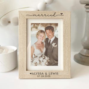 May include: A light-colored wooden picture frame with a photo of a couple. The word "married" is above the photo, and the names "Alyssa & Lewis" and the date "07.28.2025" are below. The frame is on a white surface.