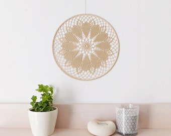 May include: A beige crocheted wall hanging with a circular design. The hanging is attached to a white wall with a string.
