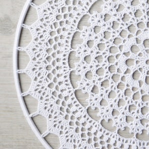 May include: White crochet doily with a circular design. The doily is made of delicate, intricate stitches and has a lacy appearance.