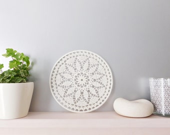 May include: White crocheted doily with a delicate floral pattern, displayed on a shelf with a potted plant and decorative objects.