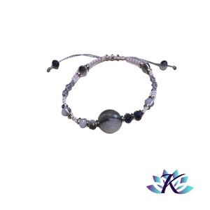 Op de afbeelding: Een wit en grijs kralenarmband met een grote grijze steenkraal in het midden. De armband is gemaakt met een wit koord en heeft kleine zwarte kralen die door de hele armband verspreid zijn.