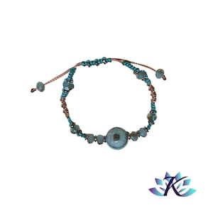 Op de afbeelding: Een turquoise macramé armband met een centrale cirkelvormige kraal en kleinere kralen. De armband heeft een bruin en blauwgroen koordontwerp, met verstelbare schuifknopen voor een aangepaste pasvorm. De armband staat op een witte achtergrond.