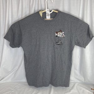 May include: A gray t-shirt with a pocket featuring a cartoon bowling design. The pocket has the text "UP YOUR ALLEY" below the bowling design. The shirt is hanging on a white hanger.