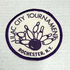 Könnte beinhalten: Ein kreisförmiger Aufnäher mit lila Rand und den Worten "LILAC CITY TOURNAMENT ROCHESTER, N.Y." in einem kreisförmigen Muster. Der Aufnäher zeigt ein lila Bowlingkugel- und Kegeldesign auf weißem Hintergrund.