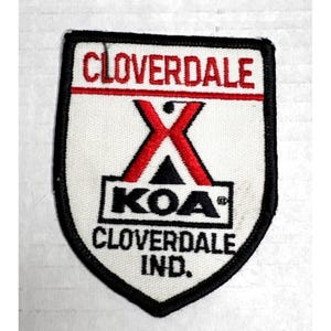 May include: A shield-shaped embroidered patch with a white background and black border. It features the red text CLOVERDALE at the top, a red and black X-shaped logo above the black text KOA, and the black text CLOVERDALE IND. at the bottom.