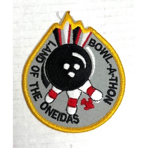 Könnte beinhalten: Grauer, gestickter Aufnäher mit Bowling-Motiv. Der Aufnäher zeigt einen schwarzen Bowlingball, rote und weiße Pins und die Schriftzüge "Land of the Oneidas" und "Bowl-A-Thon". Gelber Rand und Flammen-Design.
