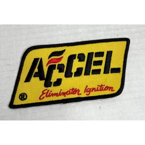 May include: A yellow embroidered patch with black and red text that reads "ACCEL Eliminator Ignition." The patch has a black border and a registered trademark symbol. The patch is rectangular with angled corners.