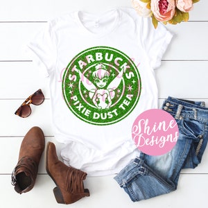 Op de afbeelding: Wit T-shirt met een groen glitterontwerp van een Starbucks-logo met Tinkerbell en de woorden "Starbucks Pixie Dust Tea".