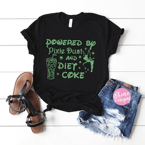 Op de afbeelding: Zwart T-shirt met groene glittertekst die "Powered by Pixie Dust and Diet Coke" luidt. Rechts van de tekst staat een groene glitterfee met vleugels. Links van de tekst staat een glas ijs met een rietje.