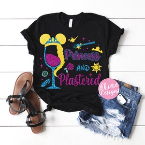 Puede incluir: Camiseta negra con un diseño brillante de una copa de vino con una corona de princesa, una varita mágica y el texto "Princess and plastered".