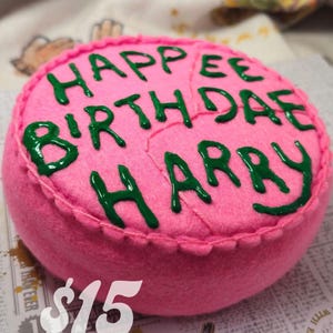 May include: A pink felt cake with green icing that reads "HAPPEE BIRTH DAE HARRY". The cake is round and has a stitched edge. The cake is a handmade item.
