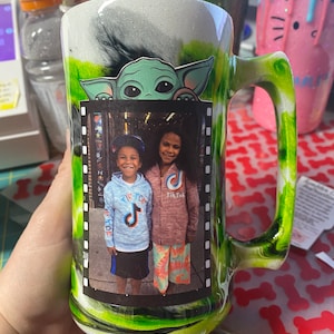 May include: A green and white ceramic mug with a black and white image of a child wearing a blue shirt with a TikTok logo and a child wearing a pink shirt with a TikTok logo. The mug has a green handle and a green and white swirl design. The mug also has a green and white image of a character from the Star Wars franchise.