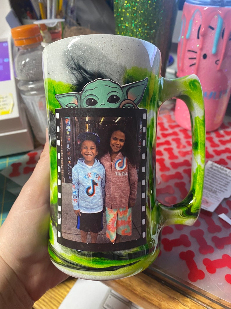 May include: A green and white ceramic mug with a black and white image of a child wearing a blue shirt with a TikTok logo and a child wearing a pink shirt with a TikTok logo. The mug has a green handle and a green and white swirl design. The mug also has a green and white image of a character from the Star Wars franchise.