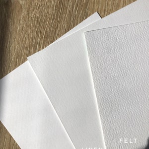 May include: Close-up of three white paper samples with different textures: smooth, linen, and felt. The paper samples are labeled with their respective textures.