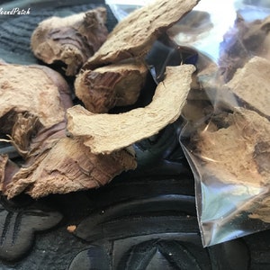 May include: Dried ginger root pieces in a clear plastic bag on a dark wooden surface. The ginger is light brown and has a rough texture.