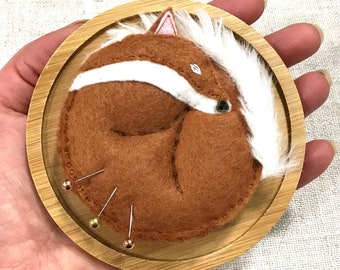 以下が含まれることがあります： 白い毛皮のアクセントが付いた茶色のフェルトのキツネの針山が丸まって眠っています。針山は、4本の金と銅のピンが突き出ている丸い木製の台座の上に置かれています。