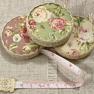 Può includere: Tre nastri di misura rotondi in tessuto con motivi floreali in tonalità di rosa, verde e crema. I nastri sono rifiniti con pizzo bianco e hanno un anello metallico in cima. Un nastro di misura bianco con segni rossi è sdraiato su una superficie di tessuto beige.