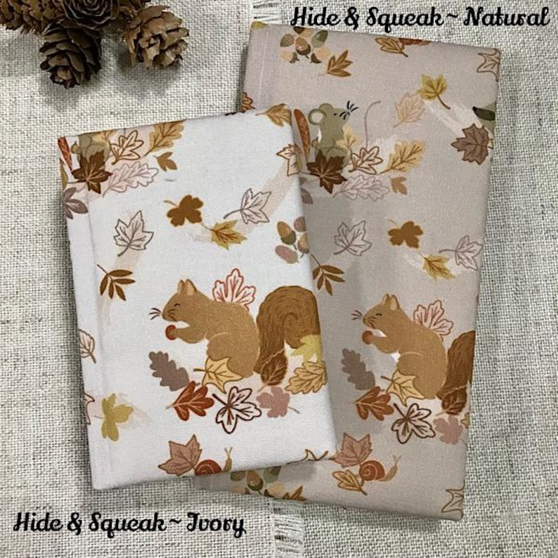 May include: Two folded kitchen towels with a fall theme. One is ivory, the other is natural. Both feature a repeating pattern of squirrels, leaves, and acorns. The text "Hide & Squeak" and "Ivory" or "Natural" are printed on the towels.