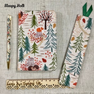 May include: A white fabric bookmark and a white fabric notebook cover with a woodland pattern. The pattern features green trees, red mushrooms, orange leaves, and a fox. The text "Sleepy Holt" is printed at the top of the image. The text "Little Bun Designs UK." is printed on a wooden ruler at the bottom of the image.