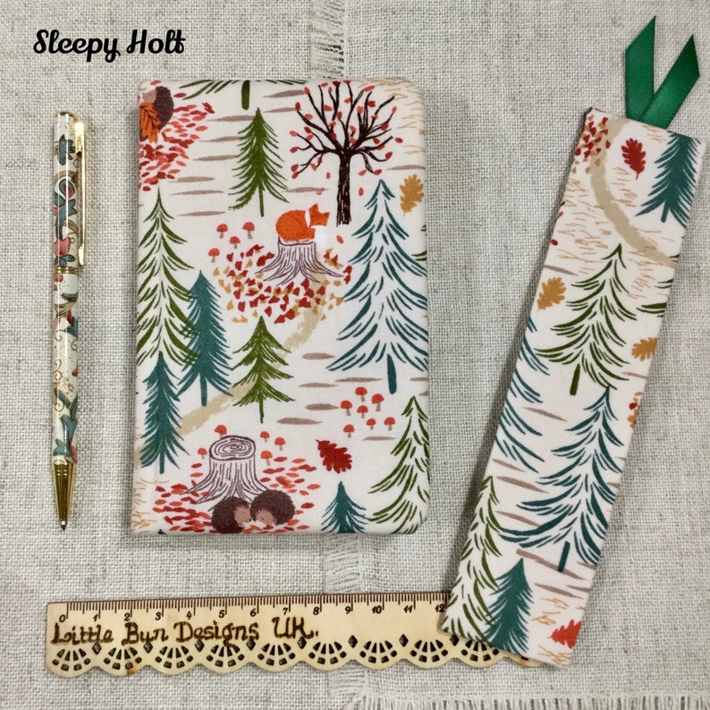 May include: A white fabric bookmark and a white fabric notebook cover with a woodland pattern. The pattern features green trees, red mushrooms, orange leaves, and a fox. The text "Sleepy Holt" is printed at the top of the image. The text "Little Bun Designs UK." is printed on a wooden ruler at the bottom of the image.