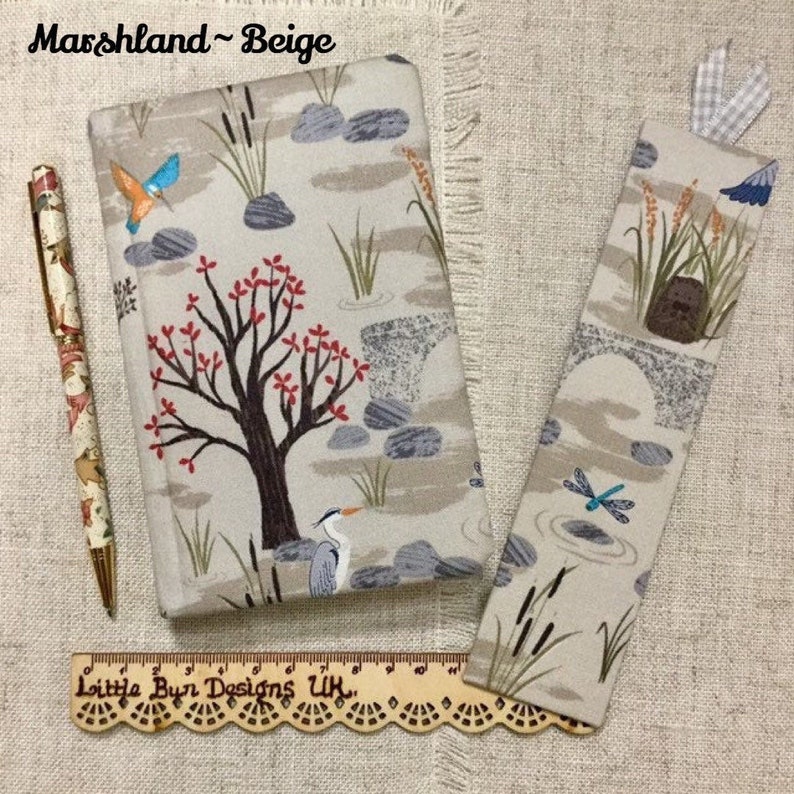 May include: A beige fabric journal with a nature-themed design featuring a tree, birds, and a heron. The journal has a matching bookmark with a dragonfly design. The text "Marshland - Beige" is printed on the cover.