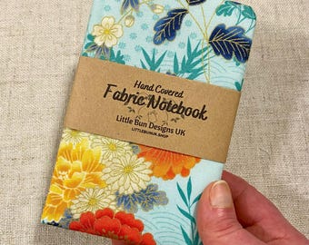 May include: A blue fabric notebook with a floral pattern and gold accents. The notebook is hand-covered and says "Hand Covered Fabric Notebook" and "Little Bun Designs UK" on the front.