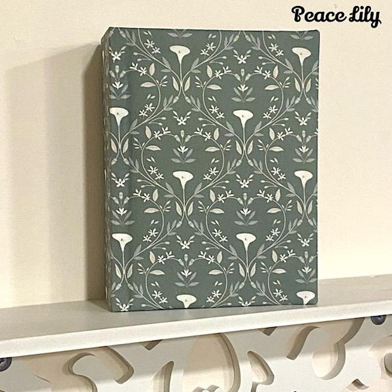 May include: A green book with a white peace lily floral pattern. The book is on a white shelf.