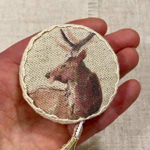 May include: A beige fabric tape measure with a brown deer printed on the front. The tape measure is round and has a gold tassel.