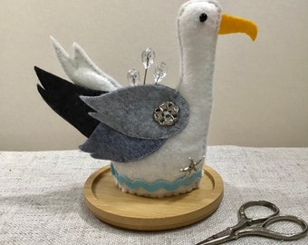以下が含まれることがあります： 白のフェルトでできたカモメの針山。黒とグレーのフェルトの羽根、青いフェルトの波の縁取り、そして星の模様が前面に施されています。針山は木製の台座の上に置かれています。